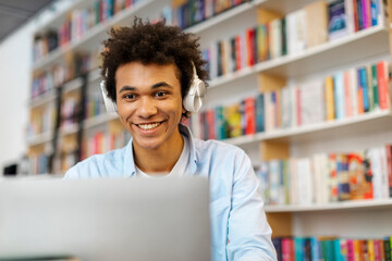 Positive Latin student guy sitting at library desk in front of laptop, enjoying online lecture via video call, free space