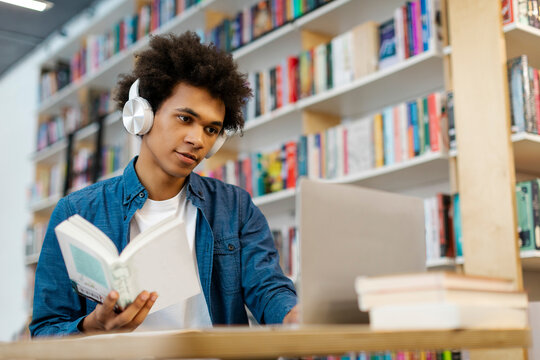Focused black male college student wearing headphone, sitting at desk in library, studying, holding book and using laptop for research or project