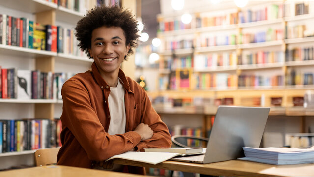 Portrait of African American smart student guy sitting at desk in university library, looking and smiling at camera