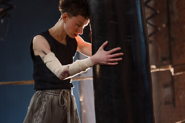 A woman in a boxing ring, tiredly leaned on a punching bag to regain her breath after an intense workout. The photo is in a cinematic style, the light gives the mood to a sports motivational story