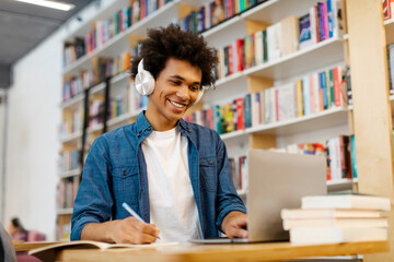 Intelligent Latin student guy sitting at desk, writing in copybook, having lecture or webinar, studying in college library. Education, new skills