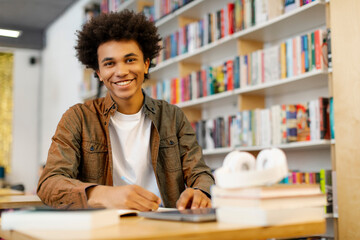 Happy intelligent African American student guy sitting at desk in library and smiling at camera while writing in copybook, free space. Exam preparation concept