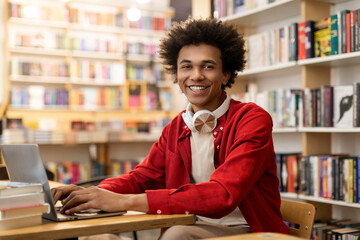 Happy African American male student sitting at desk in library smiling at camera, having online class or preparing for lecture