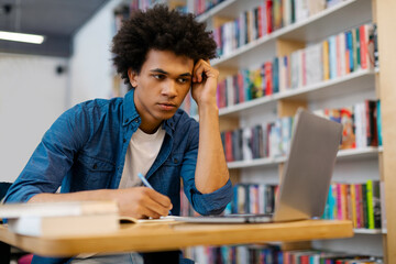 Tired focused Latin male student sitting in university library and preparing to lecture or exam, using laptop and writing notes