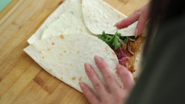 Woman assembles a shawarma wrap at home, filling a tortilla with chicken, veggies, and sauce, then rolling it for a quick homemade meal.