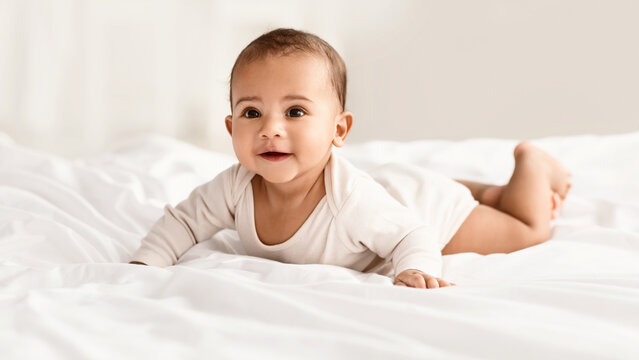 Childcare. Portrait of cute little African American baby wearing bodysuit lying on the white blanket at home. Black infant child crawling on bed in the bedroom Selective focus, blurred background - Powered by Adobe