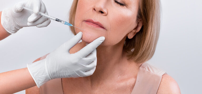 Aesthetic cosmetology concept. Senior woman getting hyaluronic acid or collagen injection on light studio background. Mature lady receiving anti-aging skincare treatment