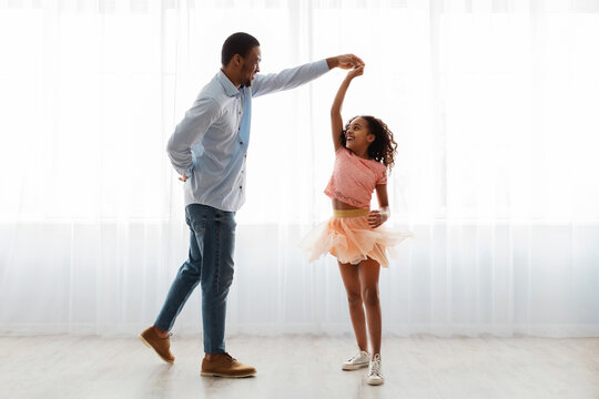 Handsome black father and little daughter in beautiful dress dancing waltz at home, standing next to window, full length photo. Loving african american dad and school girl having fun together at home
