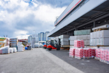 Blurred image industrial warehouse with stacks of construction materials on pallets and a red forklift. Outdoor logistics center or building supply depot with high-rise buildings in the background.