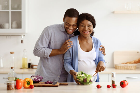Happy african american pregnant couple making dinner together. Positive expecting black woman and man cooking healthy food at home, making salad from fresh vegetables, kitchen interior