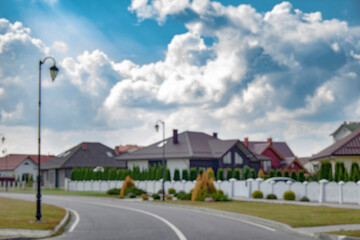 Blurred view of a quiet suburban neighborhood with private houses, white fences, and a curved street under a cloudy sky. Background for real estate, property, and urban development themes.
