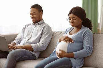 Upset pregnant african american woman sitting by her ignoring husband using laptop, chatting with...