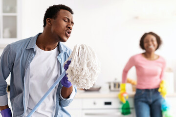 Funny black guy using mop as microphone, singing while cleaning kitchen with his beautiful...
