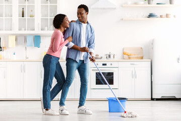 Beautiful african american young couple mopping floor in kitchen together, full length photo, copy...