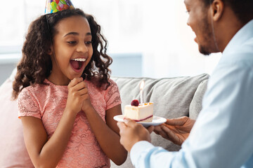 Closeup of surprised black little girl looking at cake with lit candle in her father hands, beautiful african american family dad and daughter celebrating kid birthday together at home
