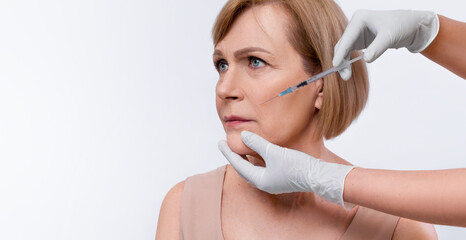 Elderly woman getting beauty nasolabial filler injection on white studio background, panorama with...