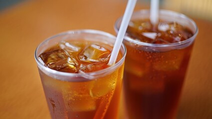 Two plastic glasses filled with iced tea, ice cubes, and white straws are placed on a wooden table, creating a refreshing summer beverage scene