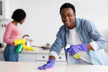 Cheerful young black guy and lady cleaning their new house together, rubbing tables with dust...