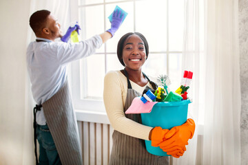 Cheerful black lady holding bucket of cleaning supplies while her husband wiping window at modern...