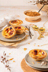 Several Pastel de Nata are arranged on plates with cinnamon, vanilla beans, and small golden spoons.
