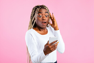 Shocked African American woman looking at smartphone in her hand, posing on pink studio background....