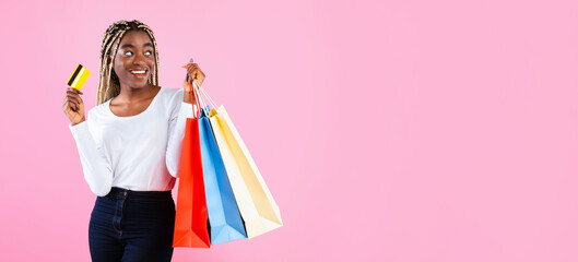 Sale And Discount Concept. Portrait of dreamy young black woman holding colorful shopper bags, thinking about gifts. Lady showing credit card and looking aside at free space, pink studio background
