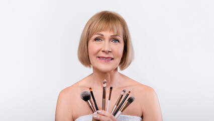 Attractive senior lady holding makeup tools pencils and brushes and smiling, posing wrapped in bath towel over light background