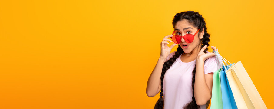 Special Offer. Portrait of surprised young indian lady wearing and touching sunglasses holding colorful shopping bags with purchases, looking at camera, yellow studio background, copy space, banner