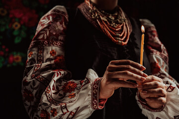 Ukrainian woman in traditional clothing holding candle in dark room interior