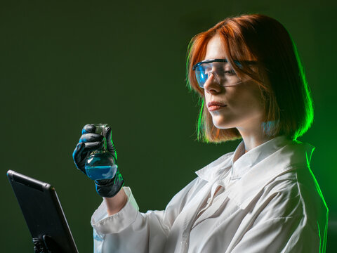 Female scientist in lab coat and safety glasses analyzing a liquid in a flask while using a tablet. Science, Chemistry, Laboratory, Research, Education, Innovation, Healthcare.