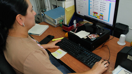 A Hispanic woman is sitting in her office at work. She is wearing a brown t-shirt and blue jeans, and she is doing some work on the computer, a task assigned by the company she works for. Next to the 