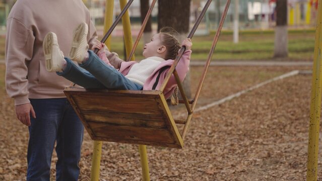 mother shakes little child on playground swing, cheerful kid flies up and down, baby laughs and smiles while playing, mother and daughter on walk in city park, happy family, childhood dream of flying.