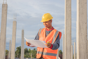Fototapeta premium Male engineer in hard hat and safety vest examines blueprint indoors at construction site; planning infrastructure project with expertise, teamwork, and efficiency.