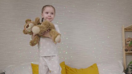 little child is jumping on bed with teddy bear in his hands, happy kid laughs in light of bright colorful lights on wall, little baby is playing with soft toy, cheerful girl in bedroom is having fun. - Powered by Adobe