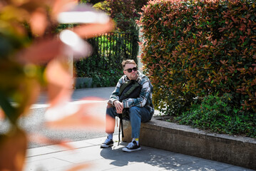 Russian male tourist resting on a stone bench near autumn shrubs in a city park, enjoying a sunny day during his travels.
