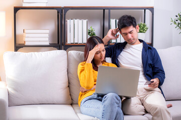 Asian couple looking laptop stressed while financial problem in living room at home, woman using laptop and man hold credit card with upset, couple with expression budget concern and expense.