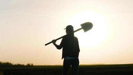 An adult girl farmer walks with a shovel on her shoulder across the field at sunset in the sky, a business lady agronomist at dawn works in the field, a man goes to drip the ground and plant seedlings