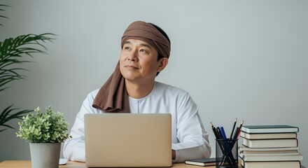 A thoughtful man wearing traditional headwear sits at his desk, using a laptop and contemplating ideas.