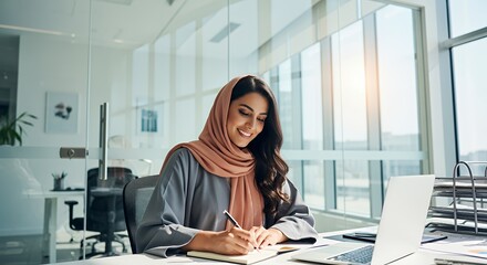 A Muslim businesswoman in a hijab works diligently at her desk in a modern , writing in a notebook and using a laptop.