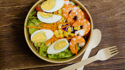 Fresh and healthy salad with shrimp, boiled eggs, lettuce, carrots, corn, and red cabbage, served in a biodegradable bowl with wooden cutlery, perfect for a light meal or takeaway