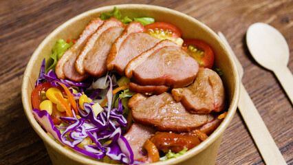 Smoked duck breast salad featuring mixed greens, tomatoes, corn, carrots, and red cabbage, drizzled with vinaigrette and served in a disposable bowl with wooden cutlery