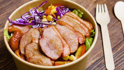 Sliced duck breast resting on a vibrant salad of mixed greens, cherry tomatoes, corn, shredded carrots, and red cabbage in a biodegradable bowl with wooden cutlery