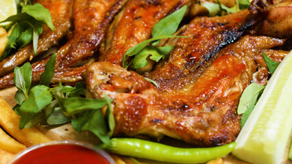 Close up of crispy roasted chicken wings seasoned with herbs, served on a wooden board with french fries, cucumber slices, a chili pepper and a small bowl of ketchup