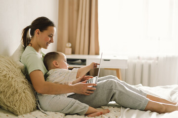 Portrait of happy young mother with little baby using laptop at home. Together, smiling mom browsing internet on computer while spending time with her little son.