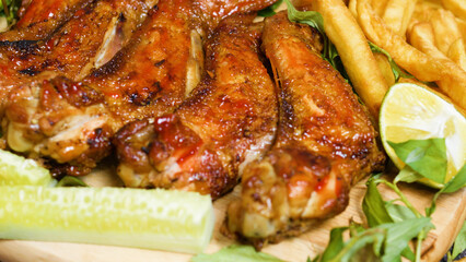 Crispy roasted chicken wings seasoned with spices and herbs, served on a wooden board alongside french fries, cucumber slices, lime wedges, and fresh herbs