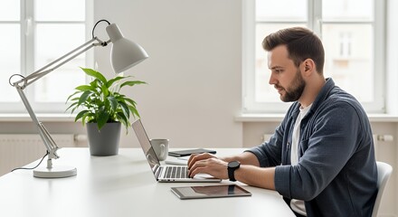A focused young man works diligently on his laptop at a bright, minimalist home desk, illuminated by a desk lamp.