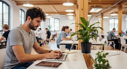 A young man works diligently on his laptop in a modern, open-plan space, surrounded by other professionals.