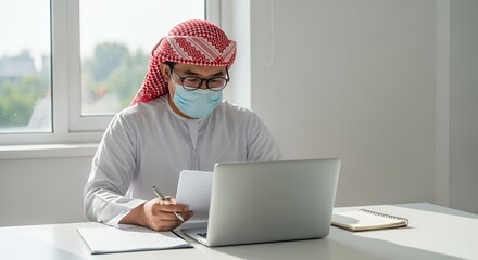 A man wearing a traditional headscarf and face mask works from home using a laptop computer.