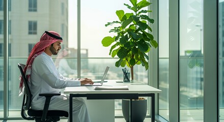 A young man in traditional Middle Eastern attire works on his laptop in a modern, bright overlooking a cityscape.