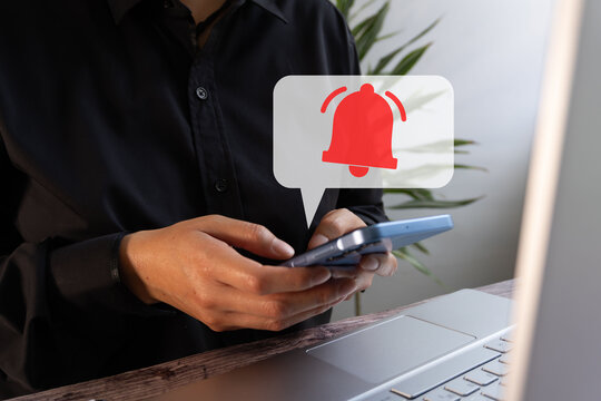 Person holding a cell phone while working in front of a computer with a virtual icon of a speech bubble and a red notification bell
